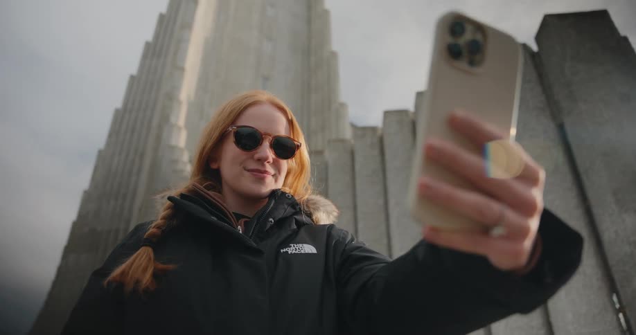 A woman with long red hair styled in a braid and wearing sunglasses and a black puffer jacket holds a smartphone at arms length to take a selfie smiling while the massive geometric structure of a landmark looms high in the background
