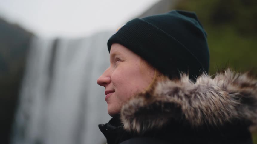 A person is shown in a closeup profile shot wearing a dark knit cap and a heavy jacket with a distinctive furtrimmed hood suggesting a cold climatethey are smiling slightly and looking offcamera with a large misty waterfall visible in the blurre
