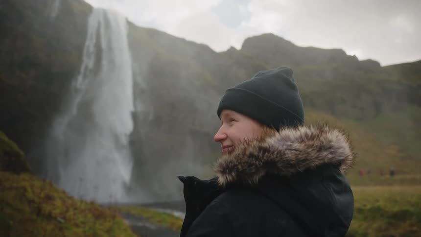 A person bundled up in a winter jacket with a furtrimmed hood and a knit cap is standing outdoors in a misty dramatic landscapethe person is looking offcamera with a slight smile and a large powerful waterfall is visible immediately behind them s