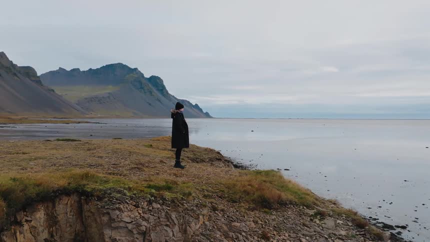 This video shows a person standing on the edge of a rugged cliff overlooking a vast calm body of waterin the background a dramatic jagged mountain range rises from the shore under an overcast sky suggesting a remote coastal landscape like iceland
