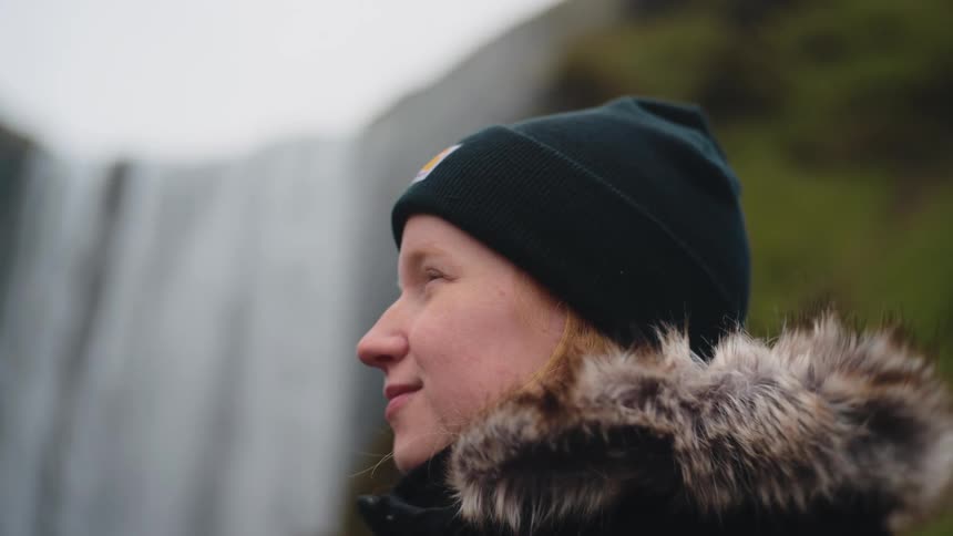 Woman in profile with dark beanie and furlined jacket observing waterfall in natural surroundings