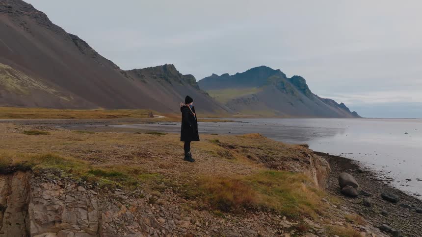 A person stands on a rocky cliff overlooking a calm bay and jagged mountains under a gray sky