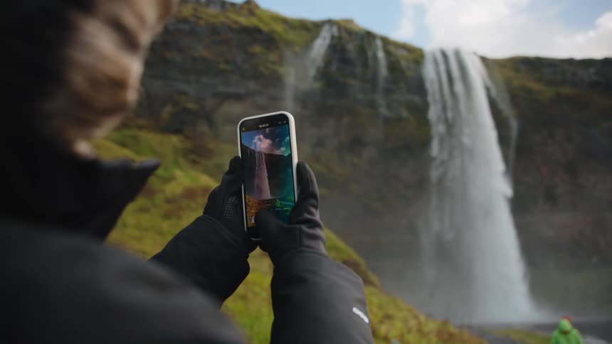 A person holds up a smartphone to photograph a waterfall in nature