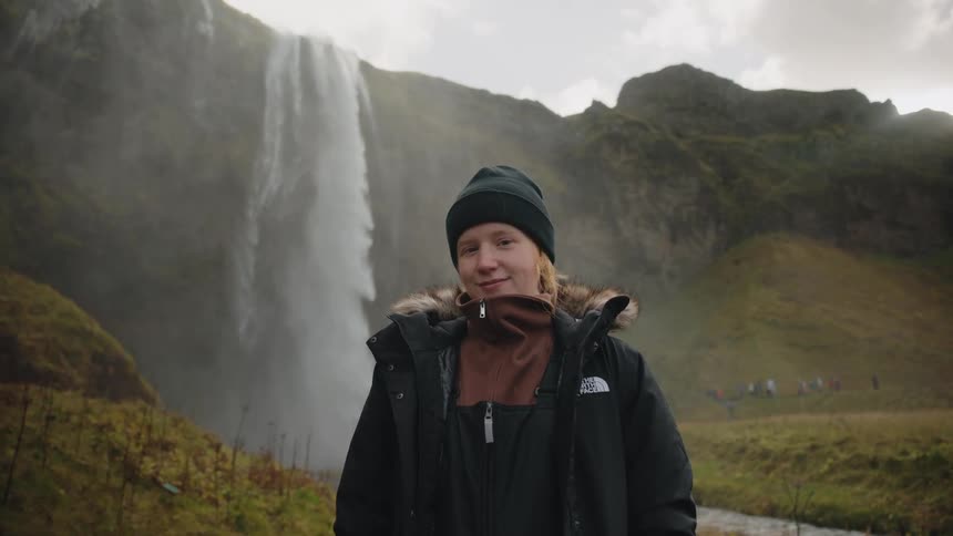A young woman wearing a black winter coat and beanie stands smiling toward the camera in front of a massive waterfall and misty green cliffs
