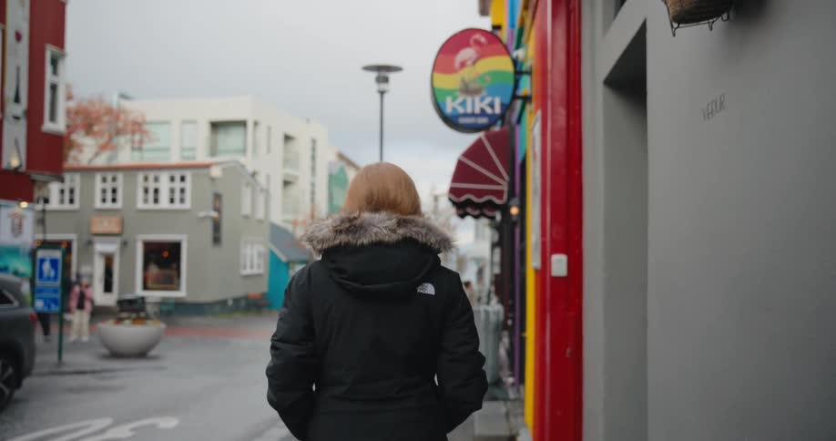 A person wearing a black winter jacket with a furlined hood walks away from the camera down a narrow wet city street on an overcast daybrightly colored storefronts including one with a rainbowcolored sign that reads kiki line the right side of t