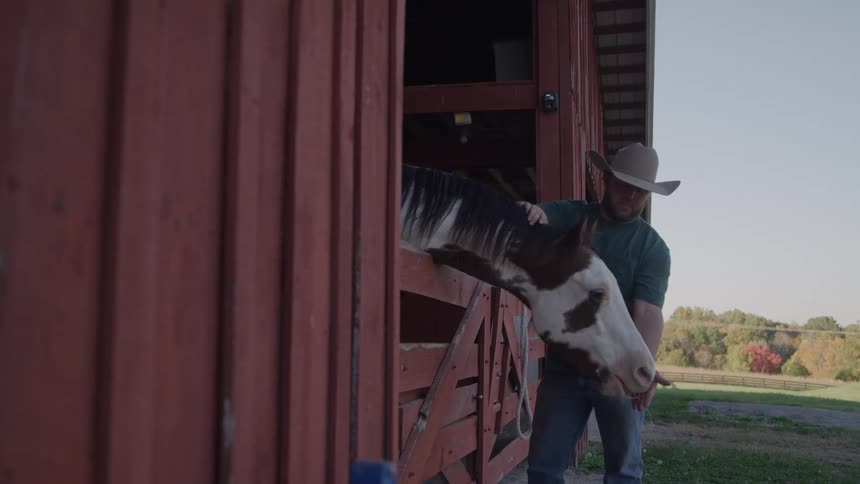 A cowboy wearing a widebrimmed hat stands in the doorway of a red barn and gently pets the head of his black and white spotted horse which pokes its head out of the stall