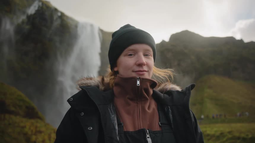 Woman poses in front of a waterfall wearing coldweather clothing and a beanie
