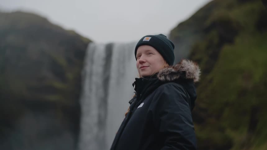 A close view shows a woman wearing a beanie and a fur trimmed parka standing near a huge misty waterfall and smiling slightly