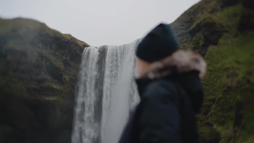 A young woman dressed in a hoodie and beanie hat watches a waterfall cascading down a mountain at midday