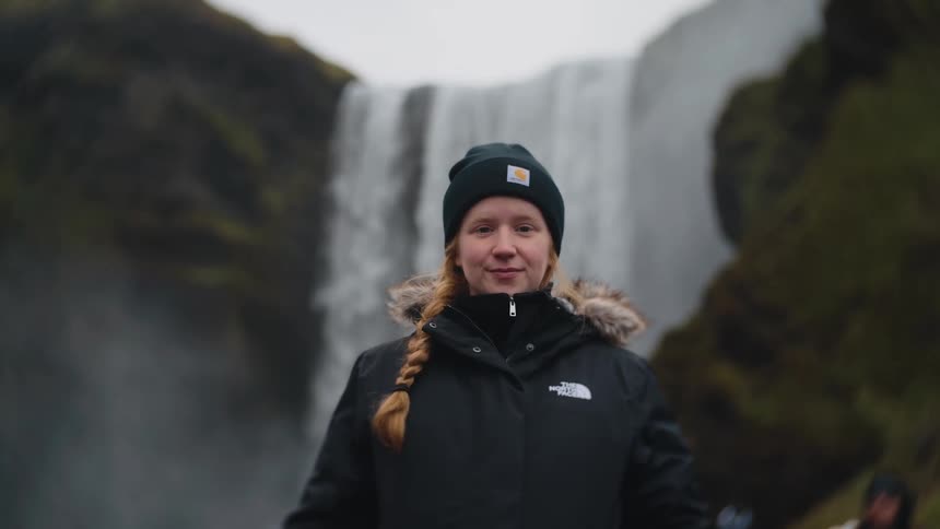 A smiling woman in a black jacket and beanie stands in front of a waterfall