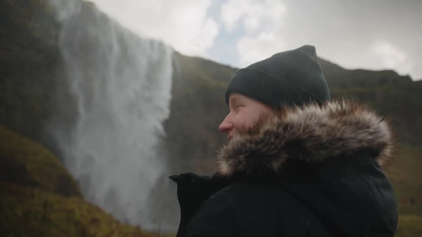 This video shows a medium closeup shot of a person possibly a woman standing outdoors and looking offcamera to the sideshe is wearing a dark possibly black winter coat with a furtrimmed hood and a matching beanie with a powerful waterfall visi