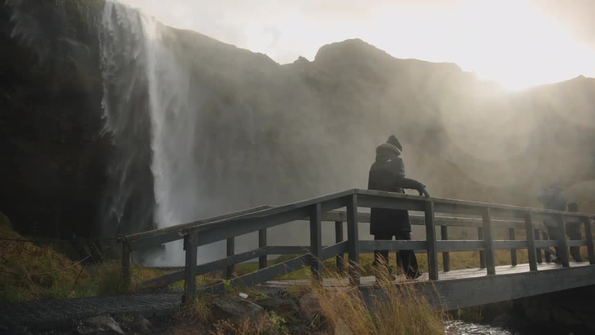 A wide view shows a person in a dark parka stopping on a wooden bridge over a stream to observe a massive backlit waterfall and misty spray