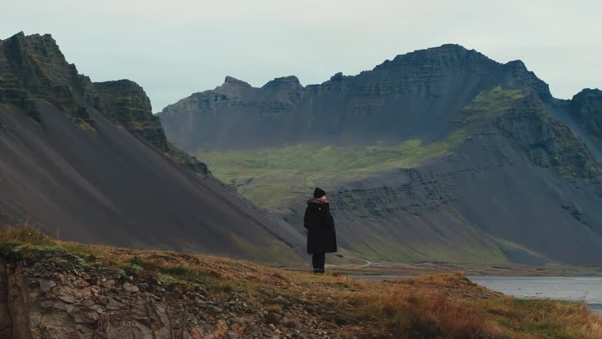 This video shows a person standing alone on a rugged grassy bluff overlooking a vast dramatic mountain landscapethe camera slowly zooms in on the person who is dressed in a dark coat and hat emphasizing the scale and solitude of the remote majest