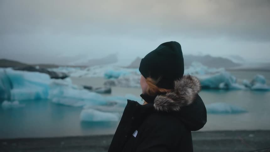 Woman in beanie and coat gazes at icebergs in icy landscape with mountains in the background