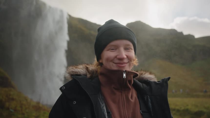 A smiling woman wearing a black beanie and a jacket with a fur trimmed hood stands in front of a powerful waterfall cascading down a cliff face