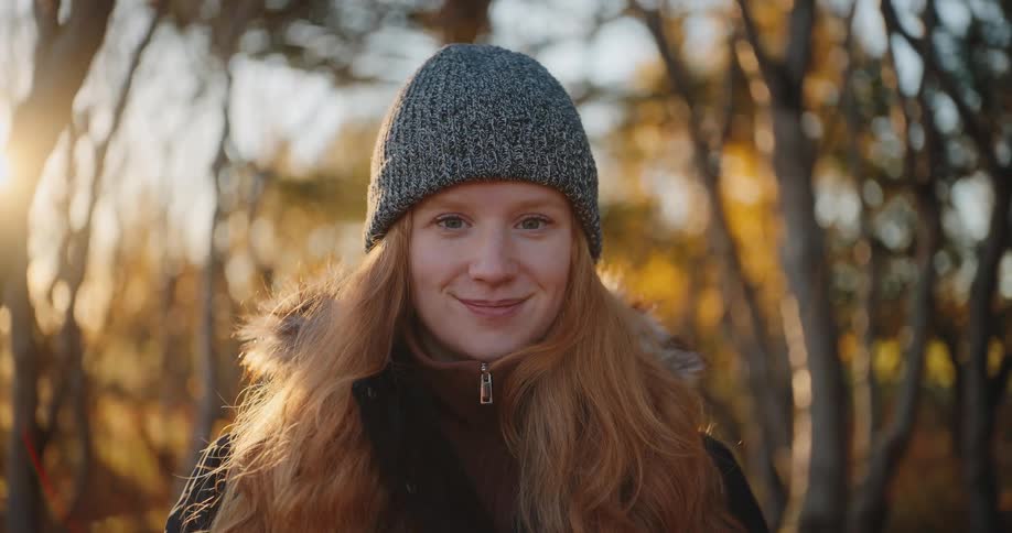 A young woman with long red hair and a grey knitted beanie wearing a dark jacket who looks down then up to make eye contact with the camera and offer a slight smile standing in a wooded area bathed in the warm soft light of the setting sun