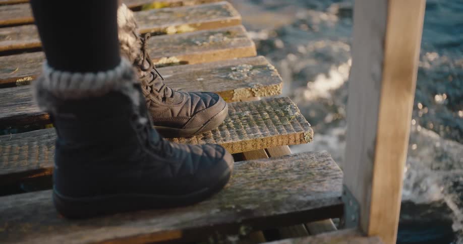 A pair of female feet in winter boots stand on a wooden bridge at midday with a river flowing beneath