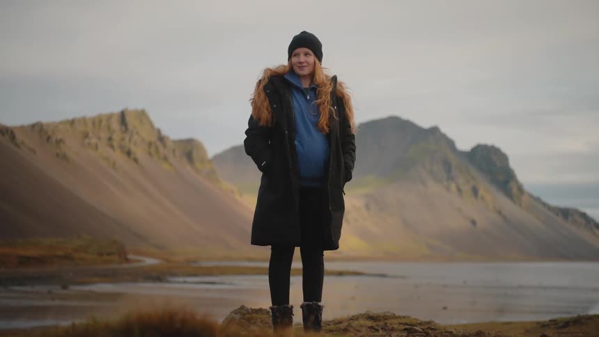 A woman with long red hair wearing a dark coat and beanie stands on a rocky outcrop overlooking a calm lake and stark mountains in a cold landscape