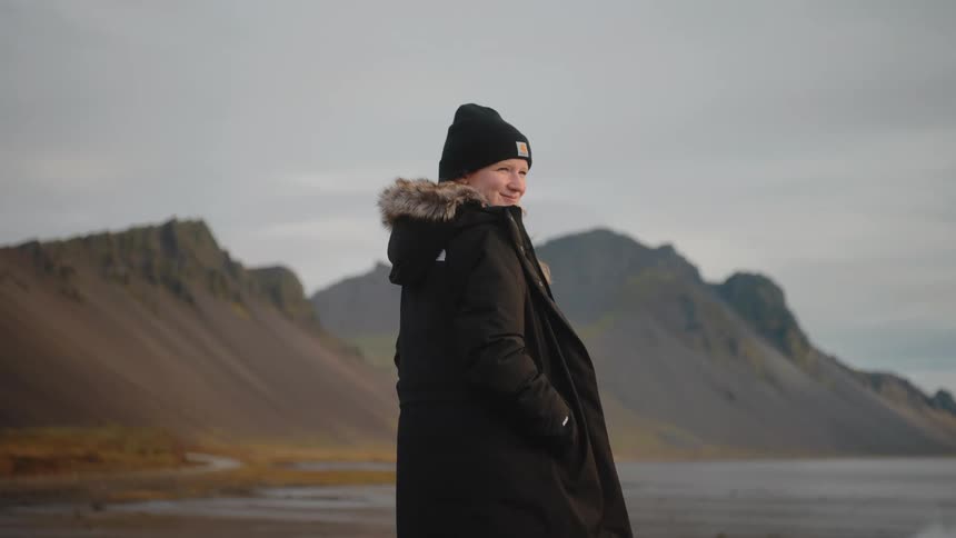 Young woman wearing a black jacket and a black beanie standing with a smiling face with dunes in the background outdoors during day time