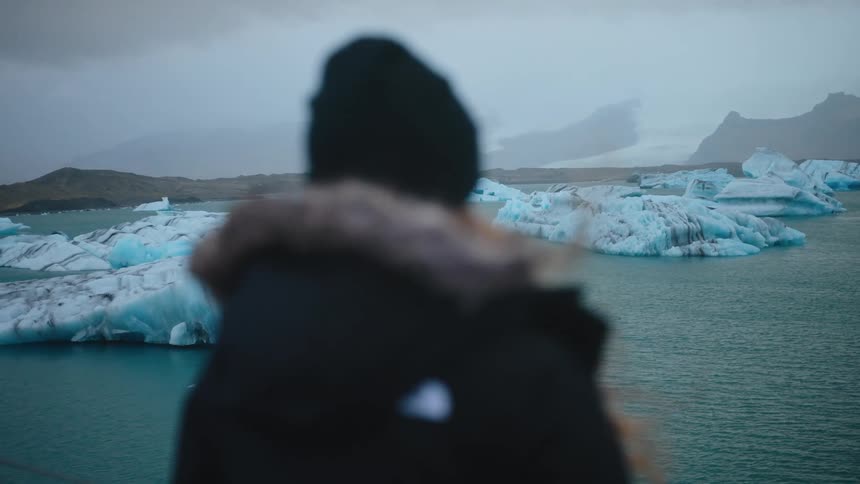 A person wearing a dark coat and hat is seen from behind looking out over a glacial lagoon filled with bright blue icebergs and surrounded by misty mountains