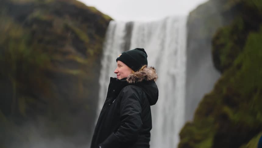 A woman wearing a black fur hooded coat and beanie smiles while looking toward a powerful waterfall cascading down a misty moss covered cliff