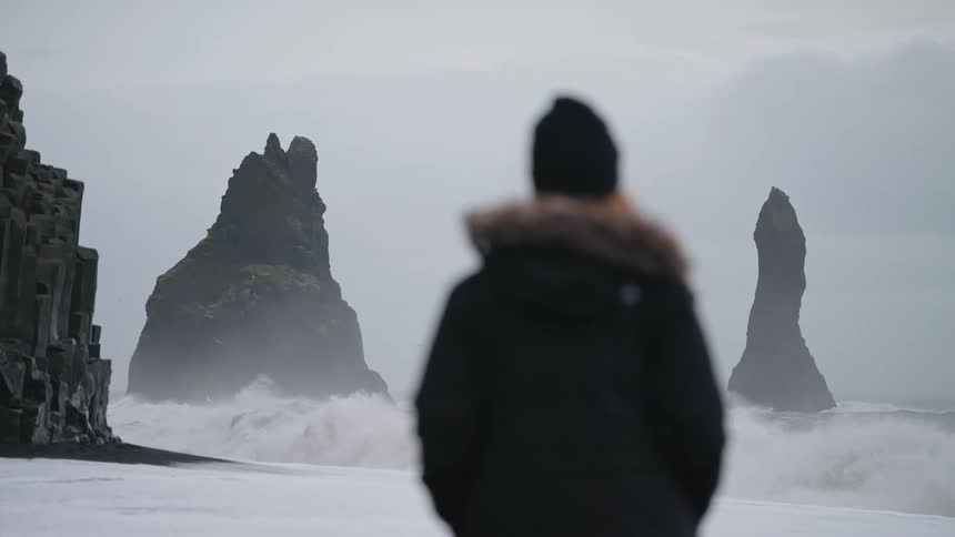 A person wearing a dark coat and hat stands out of focus in the foreground looking toward the main subject two distinct sharp basalt sea stacks rising from the ocean near a black sand beach as powerful waves crash around them under an overcast sky