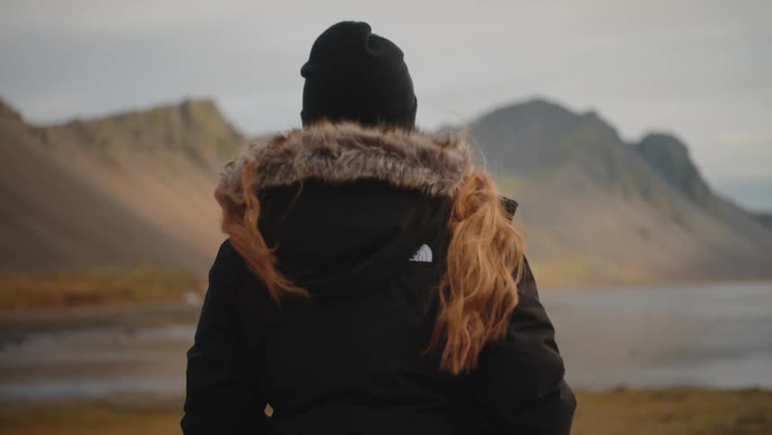 A person seen from behind stands overlooking a scenic rugged landscape with mountains and a body of water they are wearing a black winter jacket with a furlined hood and a beanie suggesting a cold environment