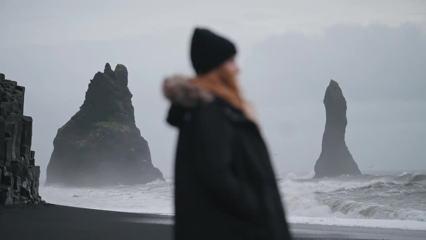 A woman bundled up in a coat and hat is on a beach in the mountains watching the waves crash on shore