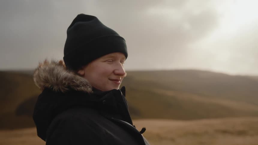 A person in a black beanie and a winter jacket with a furlined hood looks offcamera and then turns to smile directly at the camera they are standing outdoors in a remote rugged landscape with brown grasses and hills under a cloudy bright sky