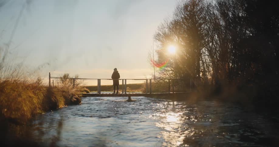A lone figure silhouetted against the bright afternoon sun stands still on a small wooden footbridge spanning a flowing river the scene is set in a natural landscape with tall dried grasses on the left and trees on the right with the sun creating a s