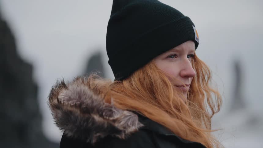 The person has long vibrant red or orange hair is wearing a black knit beanie with a small carhartt logo visible and a dark furtrimmed winter jackettheir eyes are closed briefly before they open and look intently offcamera toward the right sug