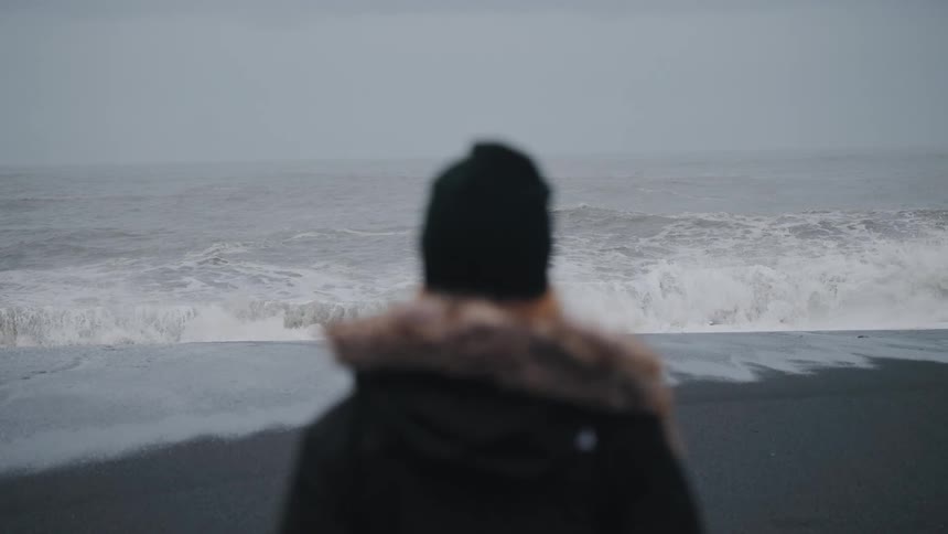 A person wearing a black beanie is standing looking out at a sea with some waves breaking on the shore