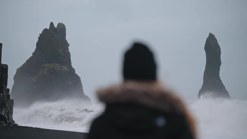 This moody shot shows a blurry figure in a winter coat standing on a black sand beach facing two distinct dramatic basalt sea stacks likely the reynisdrangar in iceland large white waves crash around the bases of the rock formations against a gray o