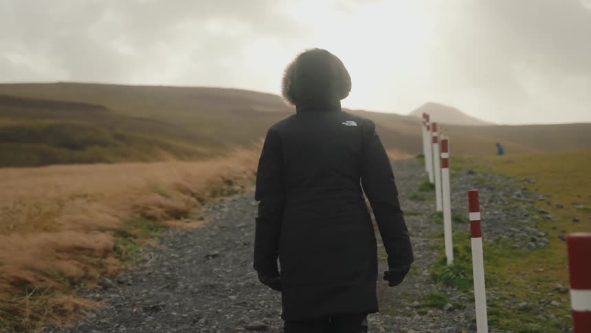 A person wearing a black hooded parka with a fur lining walks away from the camera down a gravel path surrounded by hilly rugged terrain the sky is overcast but a bright sun glare breaks through the clouds in the distance illuminating the road marker