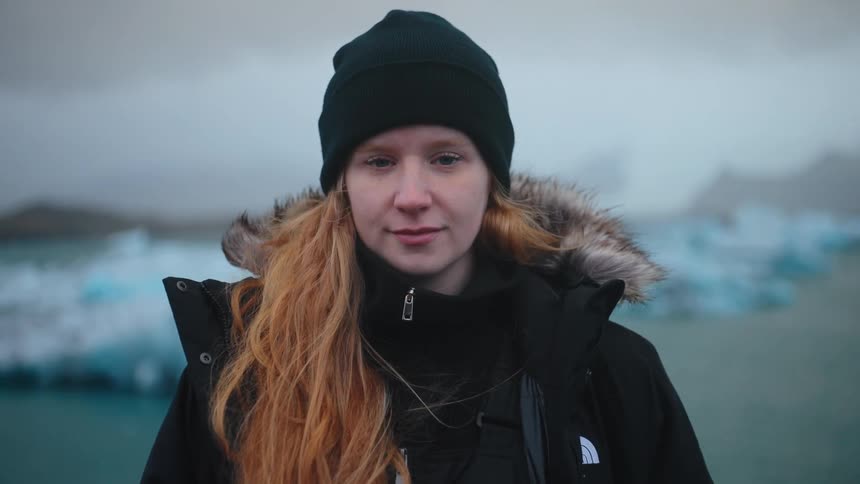 A young woman with long vibrant red hair wearing a black beanie and a warm winter jacket looks directly at the camera in a cold natural environment the background is a misty glacial lake filled with pale blue floating icebergs