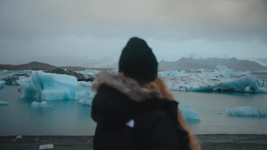 Person in beanie and furlined hooded coat watching icebergs in cold water under cloudy sky