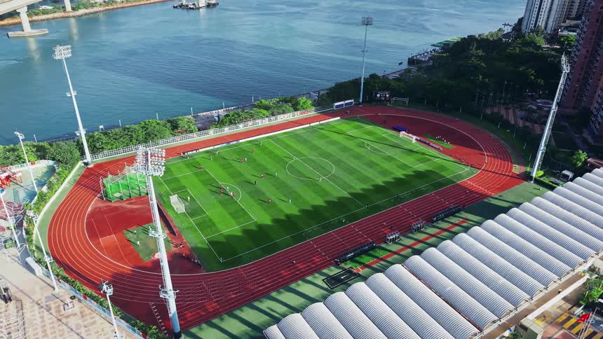 An aerial view captures a soccer match on a green field surrounded by a red running track and stadium facilities next to a waterway and tall urban buildings