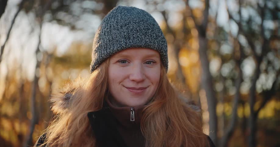 A close view shows a smiling woman wearing a gray beanie and fur trimmed jacket with long red hair illuminated by warm sunlight