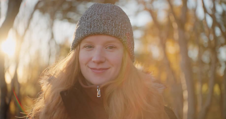 A closeup shot features a smiling woman with long red hair wearing a gray knit beanie and a winter jacket in a wooded outdoor setting the background is blurred with the warm golden sunlight creating a soft halo effect around her and a slight lens flar