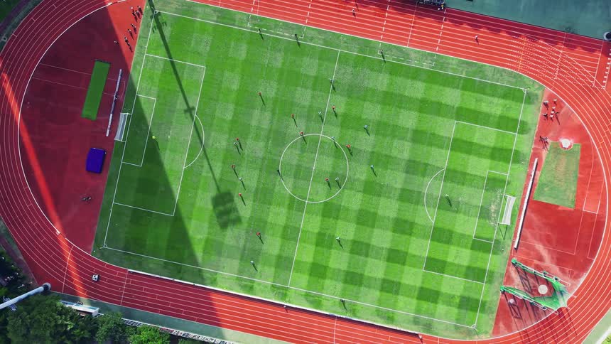 An overhead aerial view shows a soccer match in progress on a striped green grass field surrounded by a bright red running track