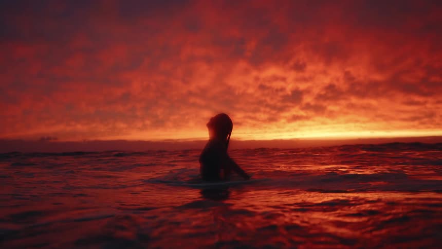 This video captures the silhouette of a person sitting in the ocean with the water around their waist or chest the scene is dominated by a dramatic sunset with intense red and orange clouds filling the sky and reflecting on the water