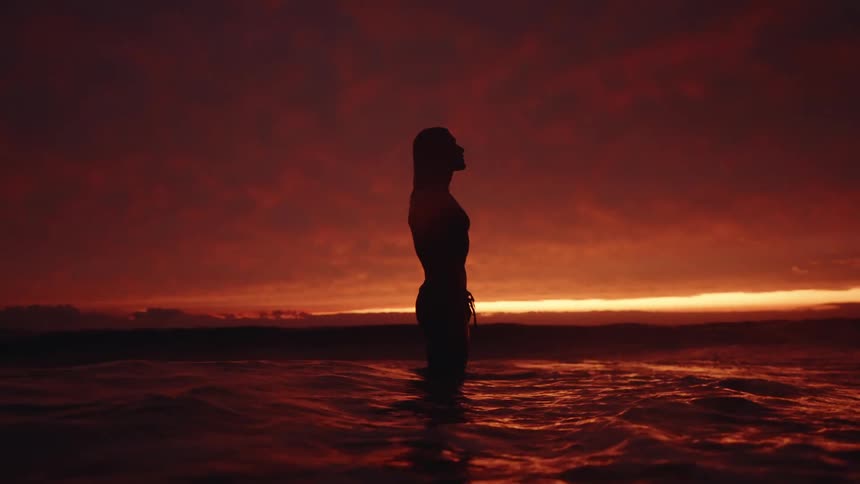 A silhouette of a woman standing in the ocean at dusk is sharply outlined against a dramatic vivid red and orange sunset sky