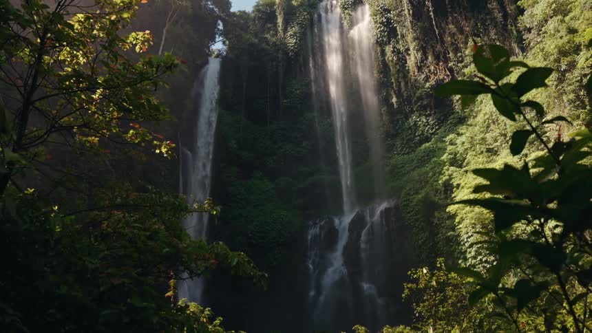 Two majestic waterfalls cascade down a tall lush green cliff face framed by dark foreground foliage in a sun drenched jungle