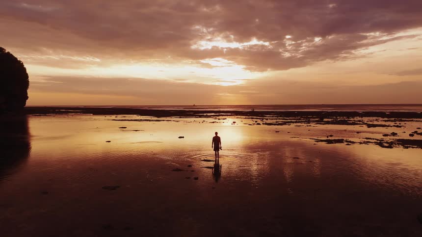 A wide view shows a person standing silhouetted in shallow water on a tidal flat beneath a dramatic cloud filled sky at sunset