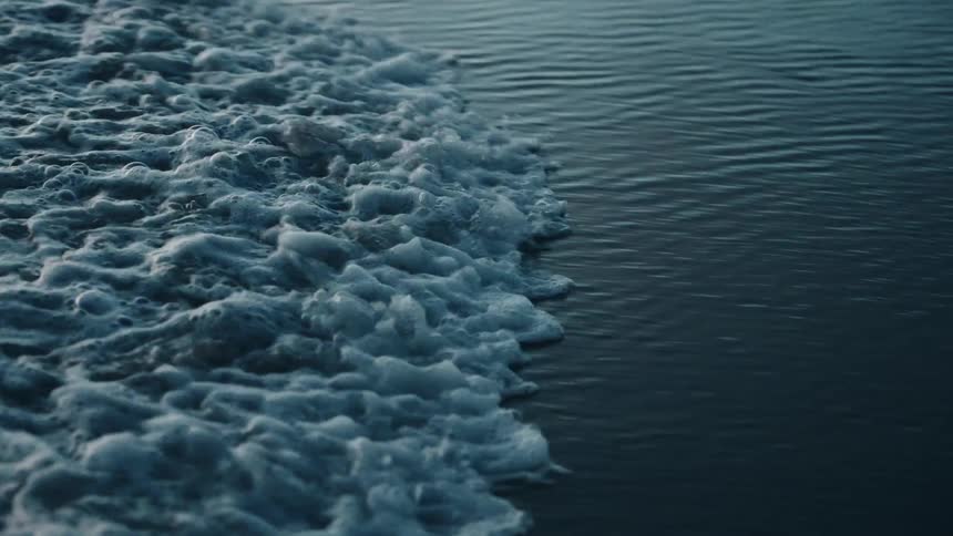 A closeup shows turbulent white froth washing up against a large expanse of deep blue calm water suggesting a wake or breaking wave