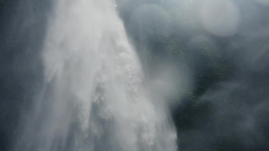 This is an extreme closeup video of a massive powerful column of waterfall plunging down the surrounding environment is obscured by a heavy mist spray and dark blurred rock or foliage