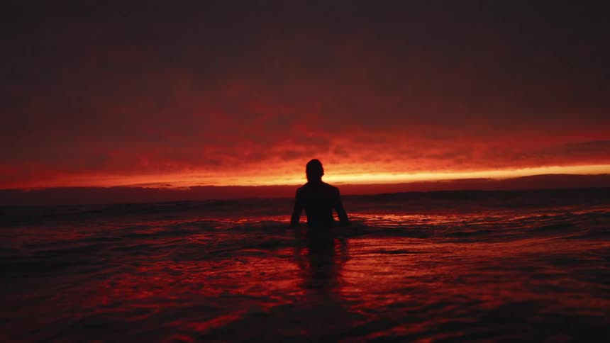 The dark silhouette of a woman emerges from the dark ocean water against a dramatic red and orange sunset sky