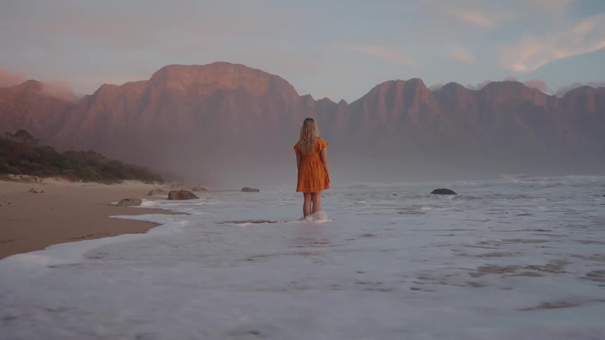 A woman with blonde hair wearing a short orange dress stands with her back to the camera on a sandy beach facing a large dramatic mountain range as the ocean waves wash around her feet during twilight hours