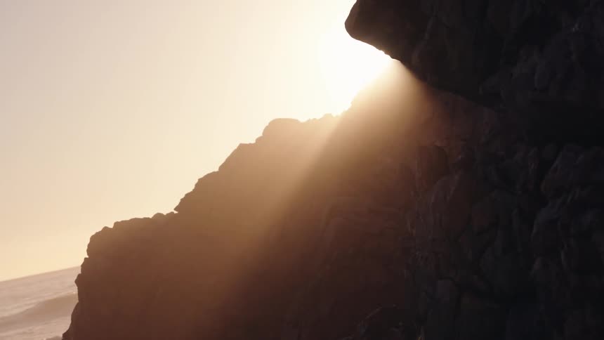 Golden sunlight flares brilliantly from behind a dark rocky cliff face by the sea