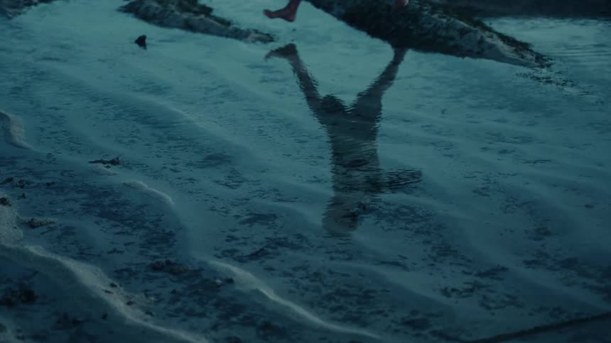 Reflection of a person appears in the shallow water as they walk along the beach